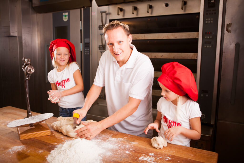 Ein Mann in der Mitte schneidet ein Brot, links und rechts von ihm stehen jeweils ein Kind, alle gemeinsam in der Backstube.