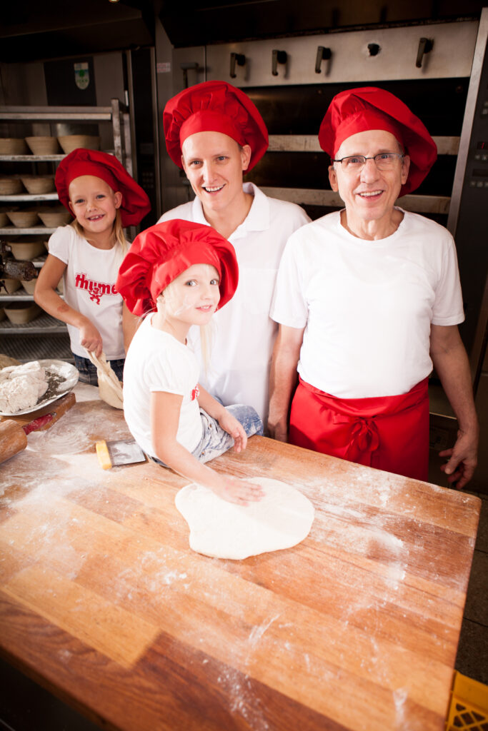 Zwei Herren und zwei Kinder in der Backstube, eines der Kinder sitzt auf dem Tisch und hält den Teig fest, während die Gruppe gemeinsam lächelt.